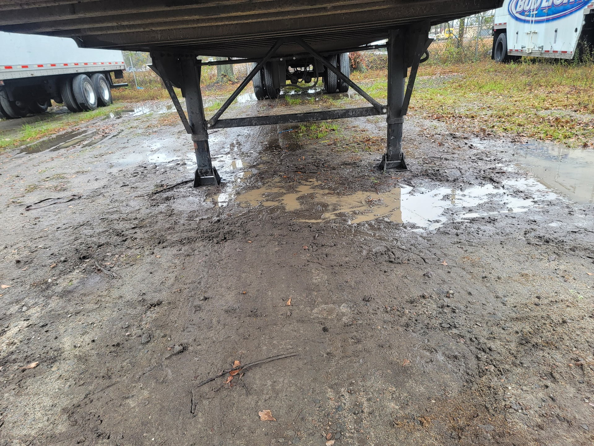 Underside of a trailer over muddy ground, with puddles and a truck visible in the background.