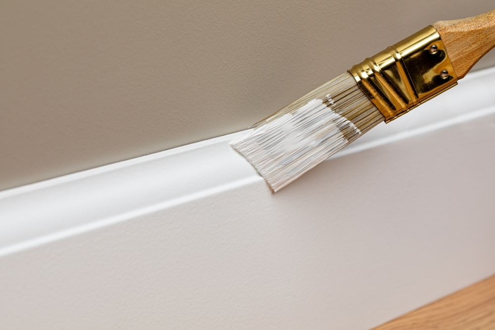 A brush is painting a white trim on a wall.