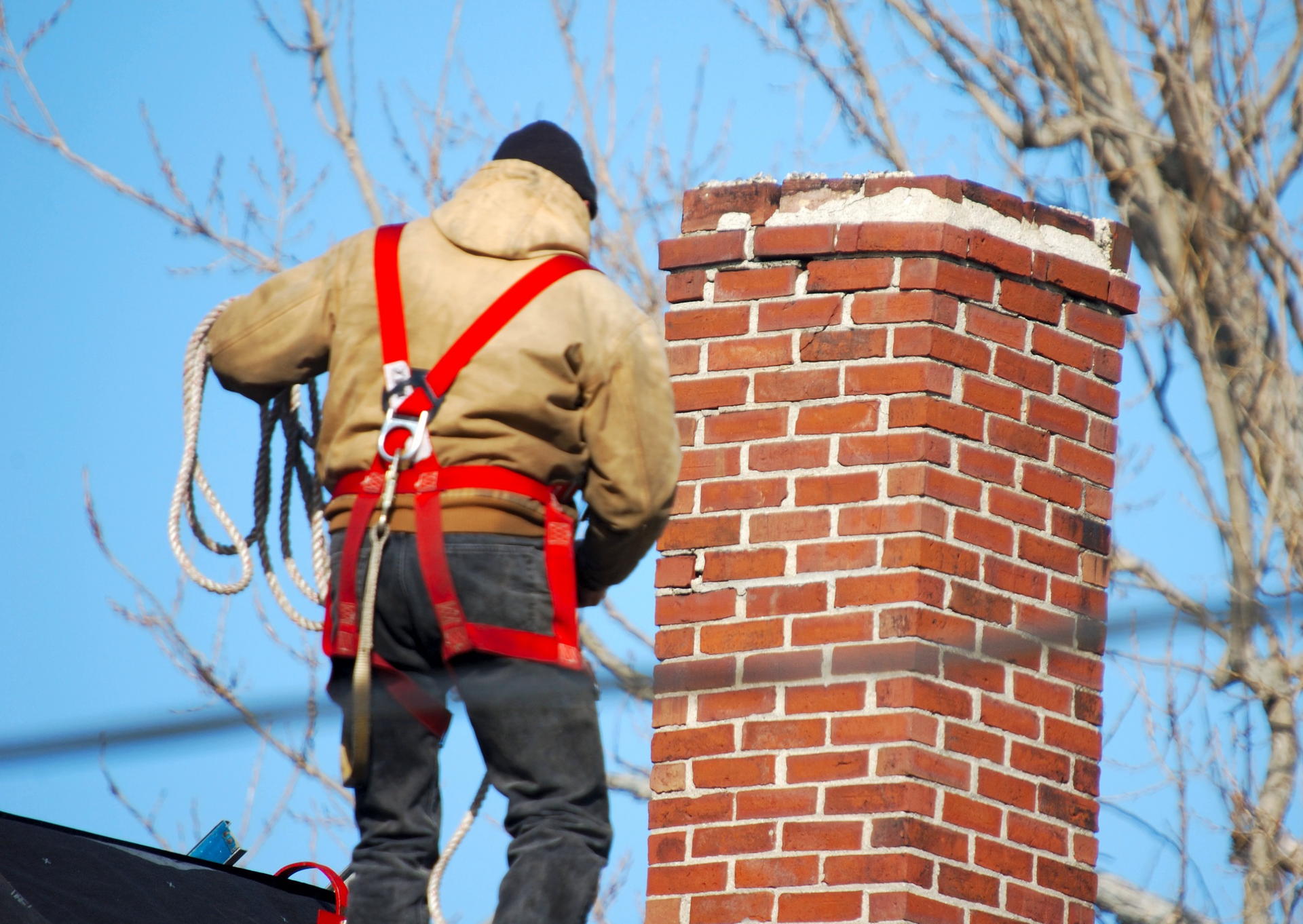 Chimney worker in harness on a rooftop, tending to a brick chimney.