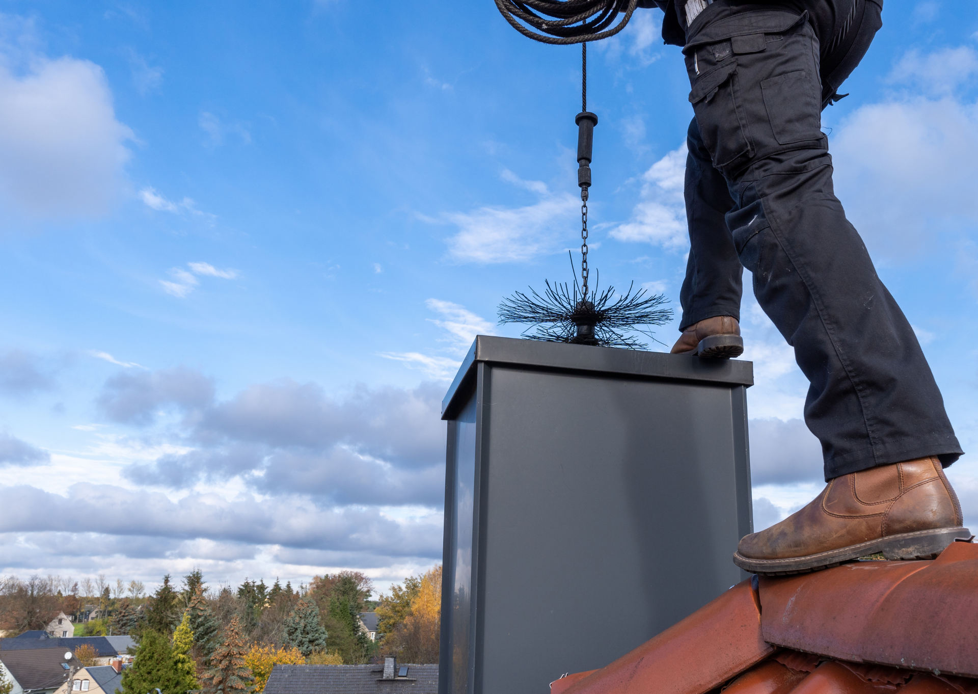 Chimney sweep on a rooftop cleans a chimney, using a brush, under a cloudy sky.