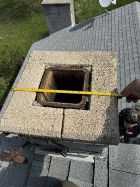 Measuring the top of a chimney on a gray shingled roof; a person's arm holds a measuring tape.