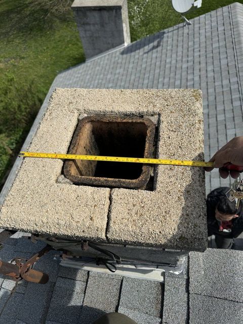 Measuring the top of a chimney on a gray shingled roof; a person's arm holds a measuring tape.