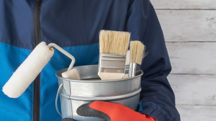 A person is holding a bucket of paint and paint rollers.