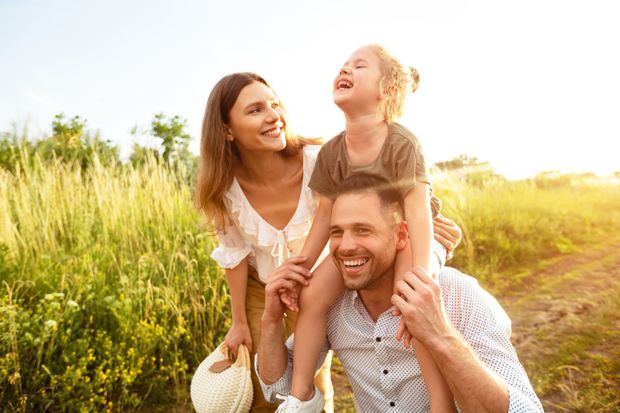 Family laughing outdoors in a sunny field, with a child on a man's shoulders and a woman beside them
