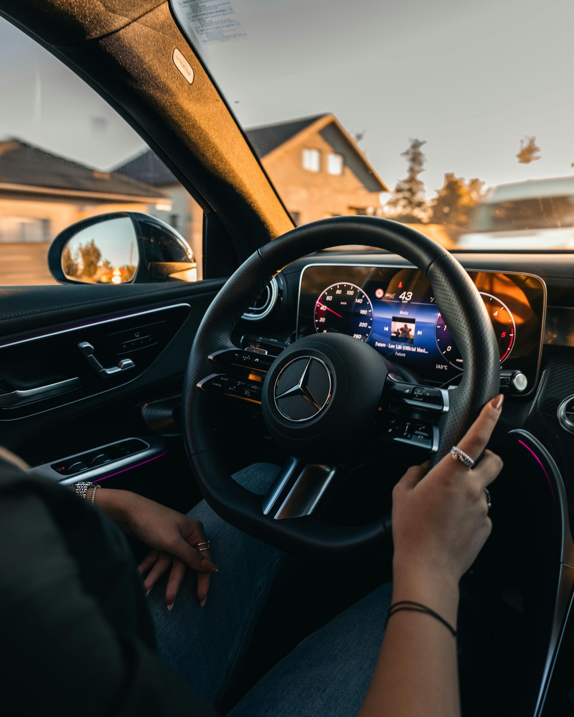 Mercedes steering wheel and dashboard seen from inside a car at sunset, with a hand on the wheel.