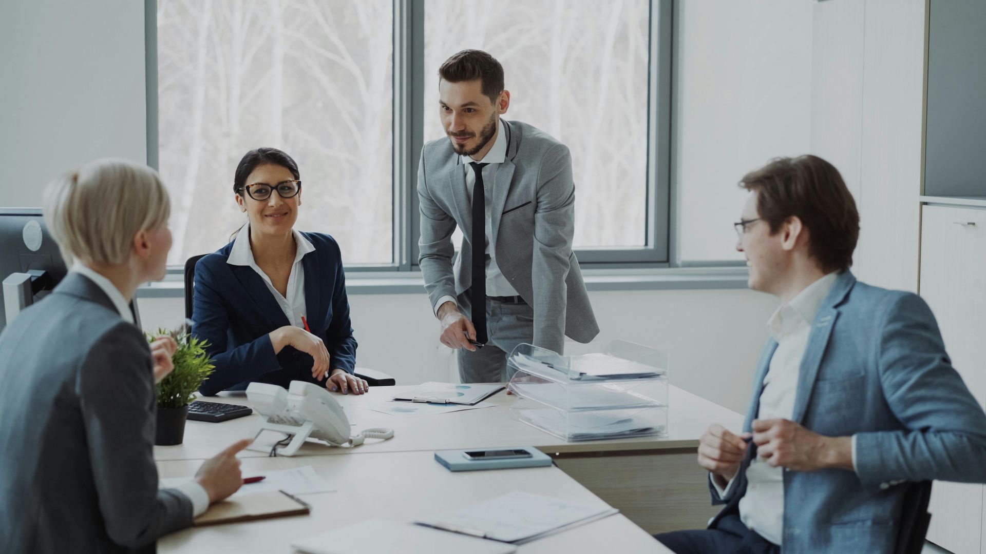 Business team meeting around a conference table in a bright office, with one man standing and speaking.