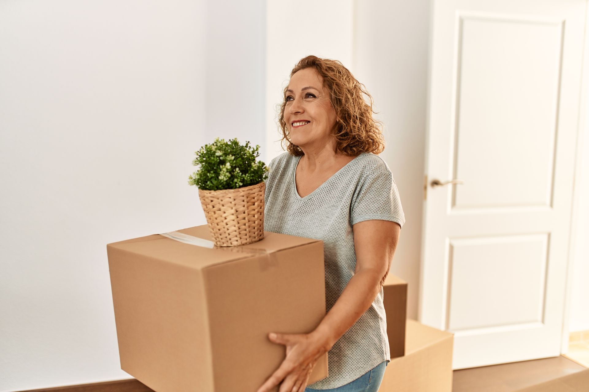 Woman holding a box and a plant, smiling. Inside a new home, near a white door and boxes.