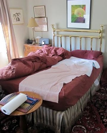 Bedroom with unmade bed, brass headboard, maroon bedding, small table with paper towel roll.