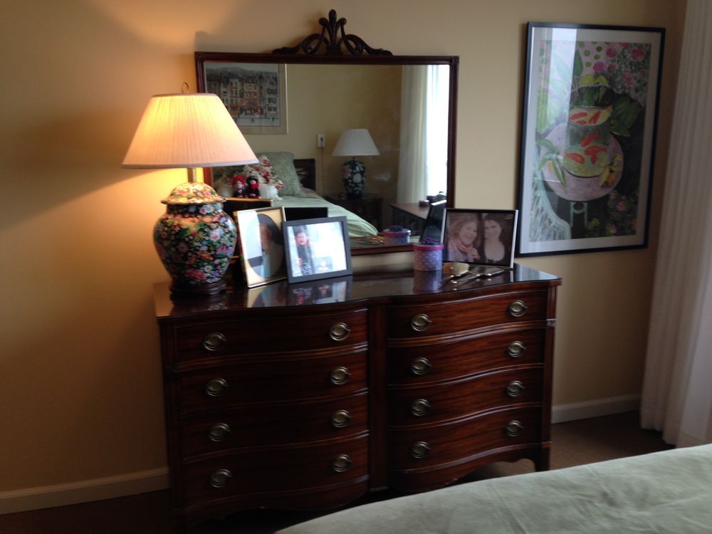 Wooden dresser with mirror and lamp, framed art on wall, a bed in view.