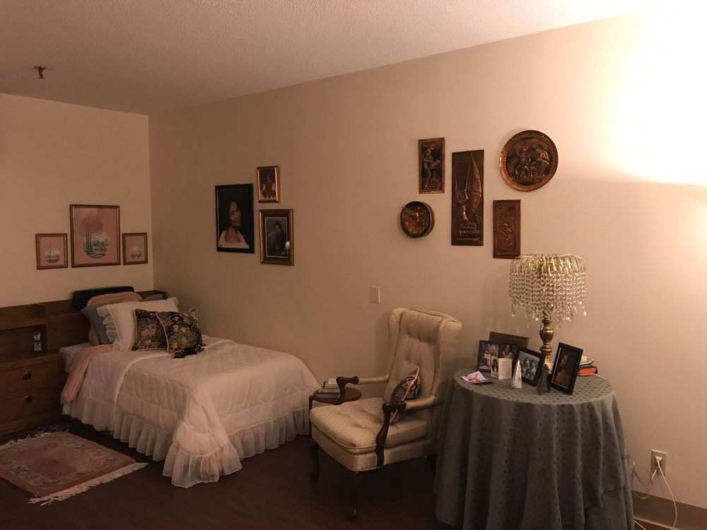 Cozy bedroom with bed, chair, side table. Wall art, lamp, and framed photos. Warm lighting.