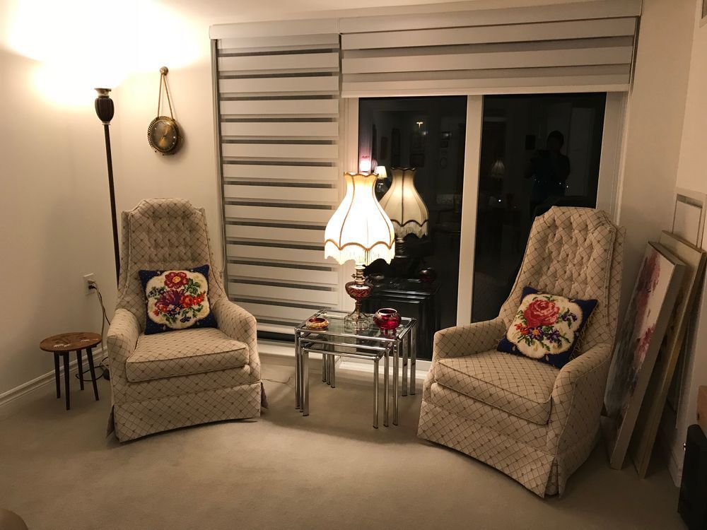 Two patterned armchairs face a window, with a side table, lamp, and floor lamp in a cozy room.