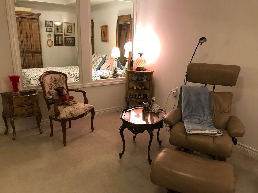 A room with a chair, ottoman, side table, mirror reflecting a bed, and another chair.