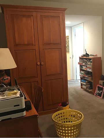 Wooden wardrobe in a room with a desk, bookshelf, and laundry basket. An open doorway is in the background.