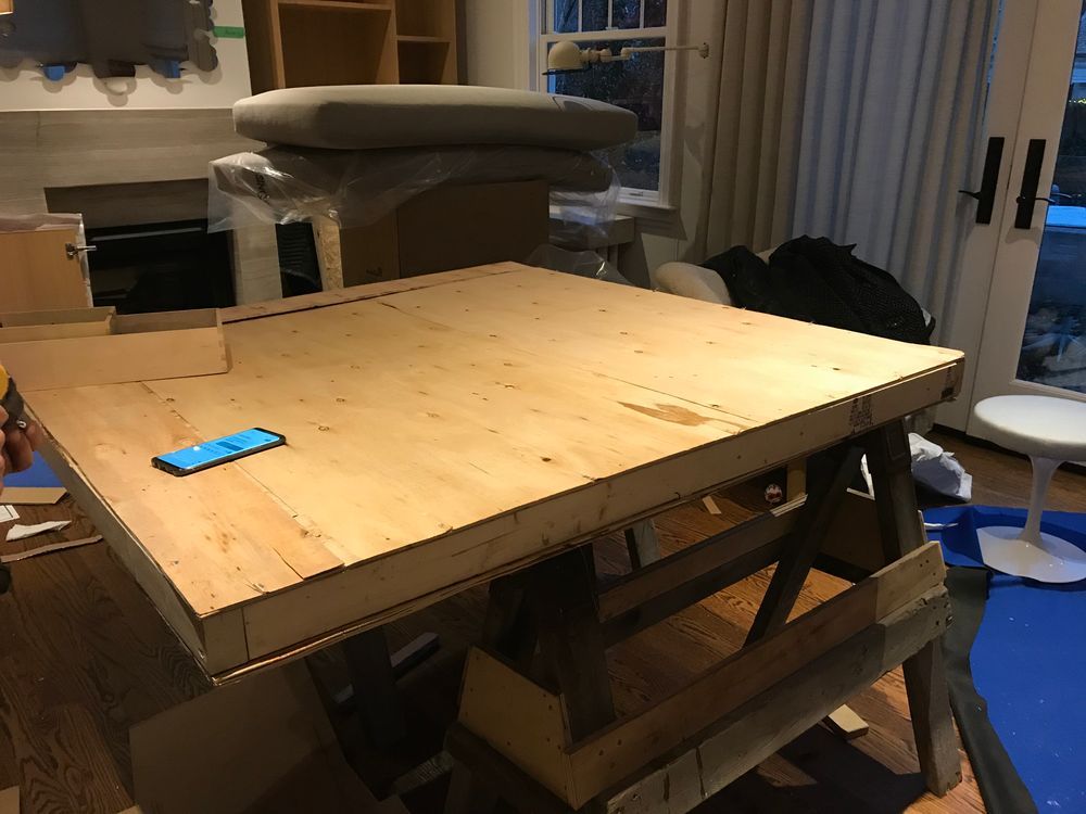 A large wooden table on sawhorses in a room with a fireplace and windows. A cell phone sits on the table.