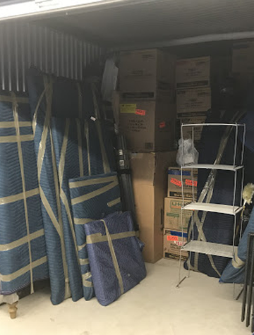 Storage unit packed with furniture wrapped in blue pads, boxes, and a metal shelving unit.