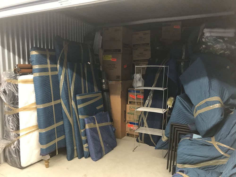 Storage unit packed with furniture wrapped in blue pads, boxes, and a metal shelf.