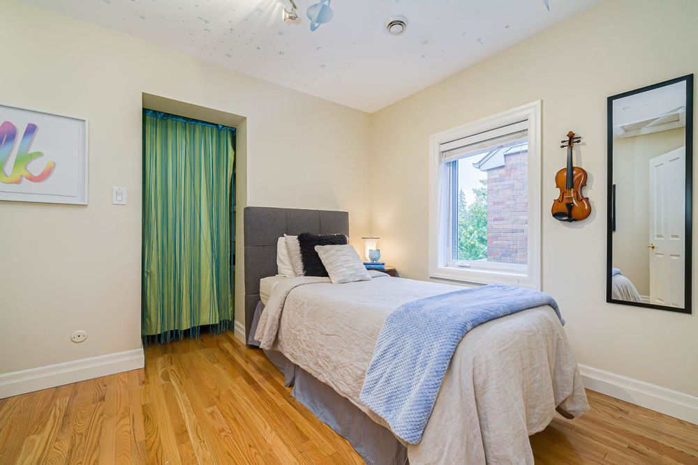 Bedroom with bed, window, violin, and full-length mirror. Green curtain in doorway. Beige walls, wood floors.