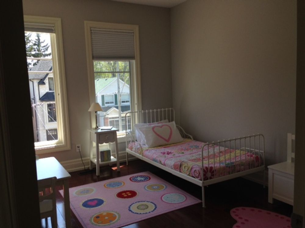 Child's bedroom with a white bed, pink rug, and two windows with a view of houses.