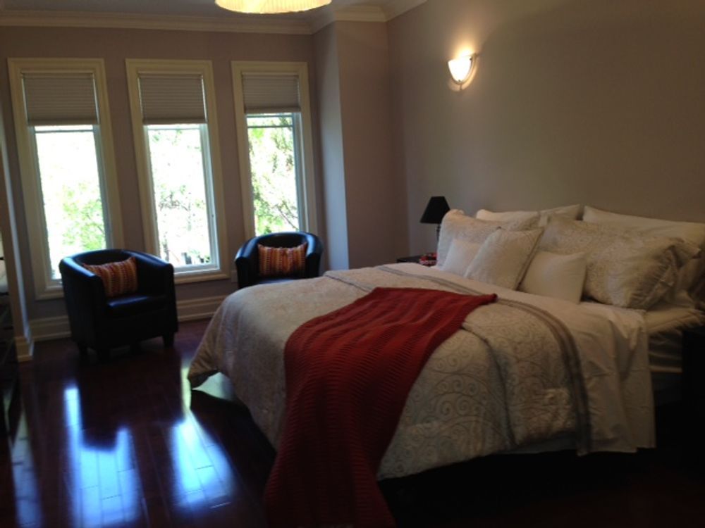 Bedroom with bed, two chairs, three windows, and dark wood floors.