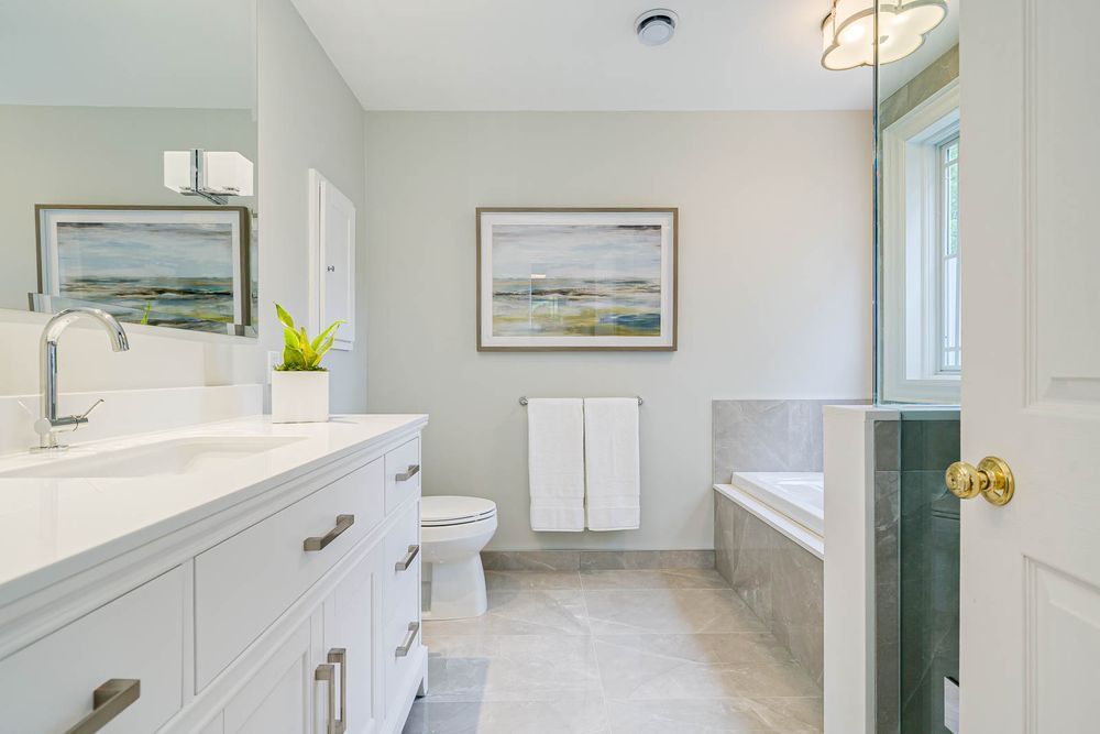 White bathroom with a vanity, toilet, artwork, and a tub with glass enclosure.