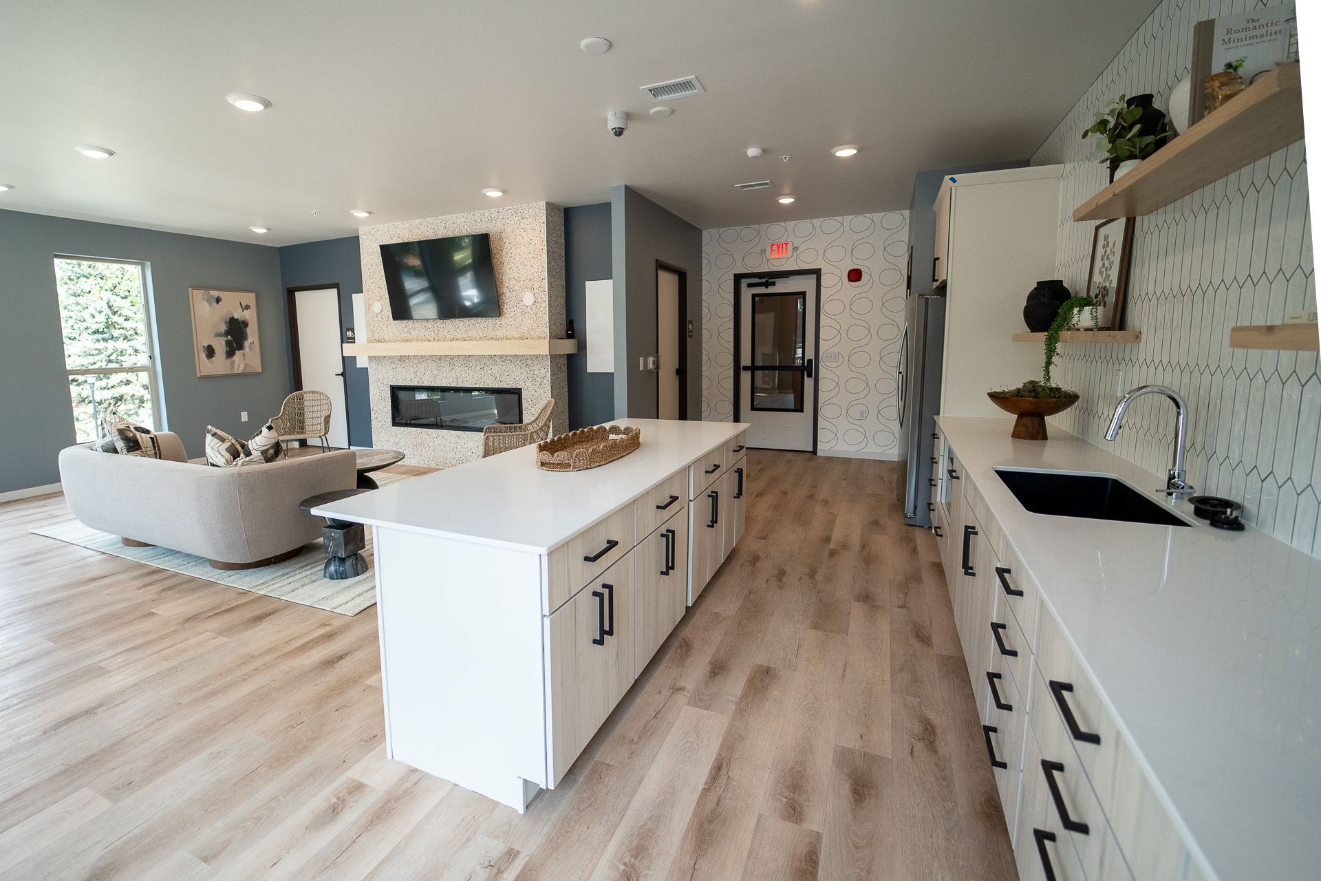 Modern open-concept community room and kitchen with light wood floors, white cabinets, a sleek counter, cozy sofa, fireplace, and wall-mounted TV. Calm and inviting space.