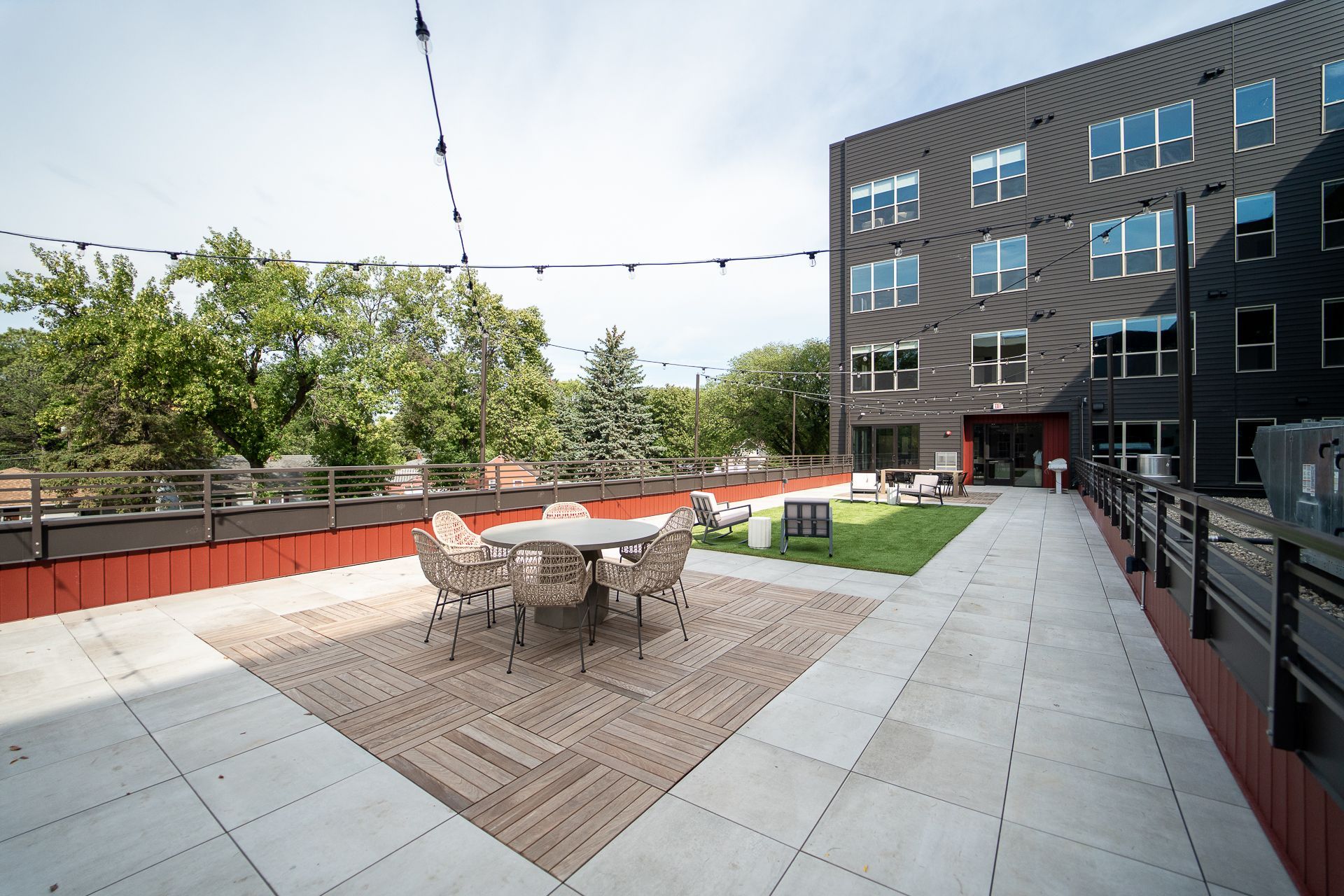 Rooftop patio with a wooden deck and wicker chairs around a table. String lights hang overhead. Adjacent modern building and trees in the background.