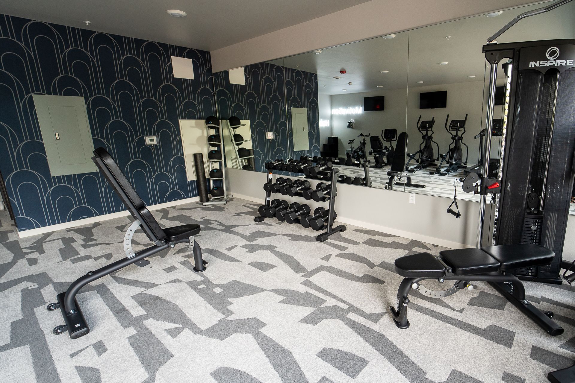 A modern gym with mirrored walls, featuring exercise machines, a bench, and dumbbells on racks. The atmosphere is sleek and energizing.