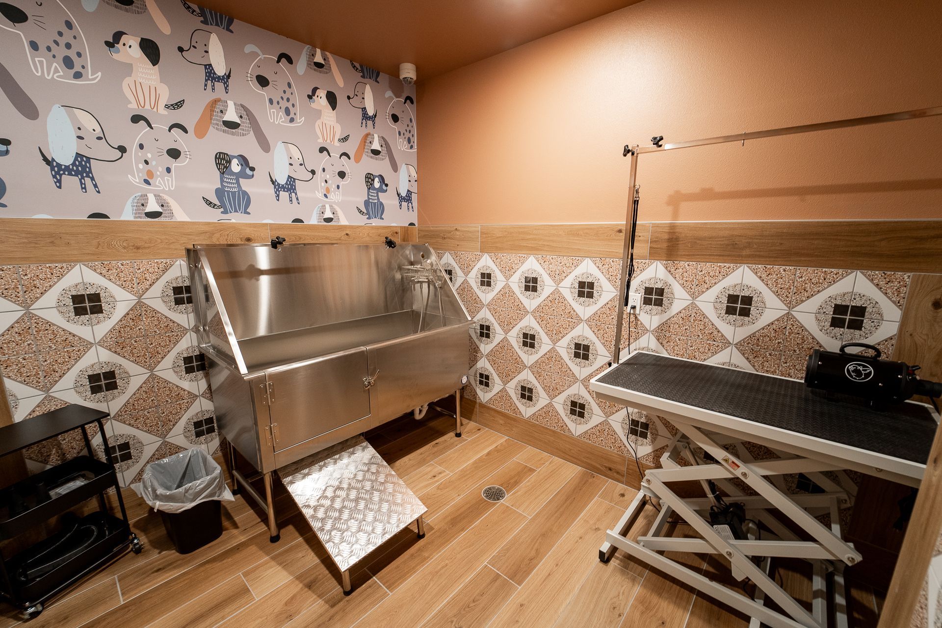 A pet grooming room with a stainless steel wash tub, dog-themed wallpaper, brown and white tiled walls, a grooming table, and a wood floor. Cozy ambiance.
