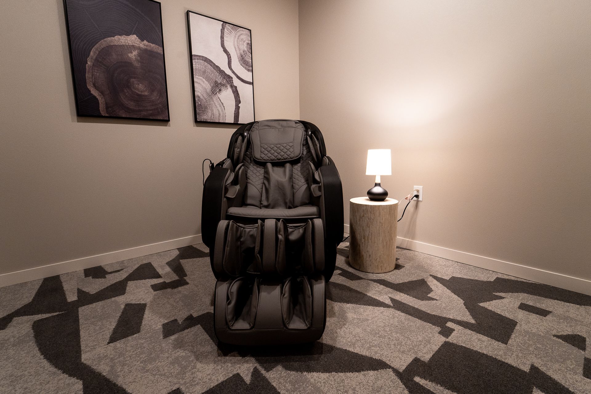 Massage chair in a cozy room with geometric carpet, two abstract artworks on the wall, and a small wooden table with a lit lamp, creating a calm ambiance.