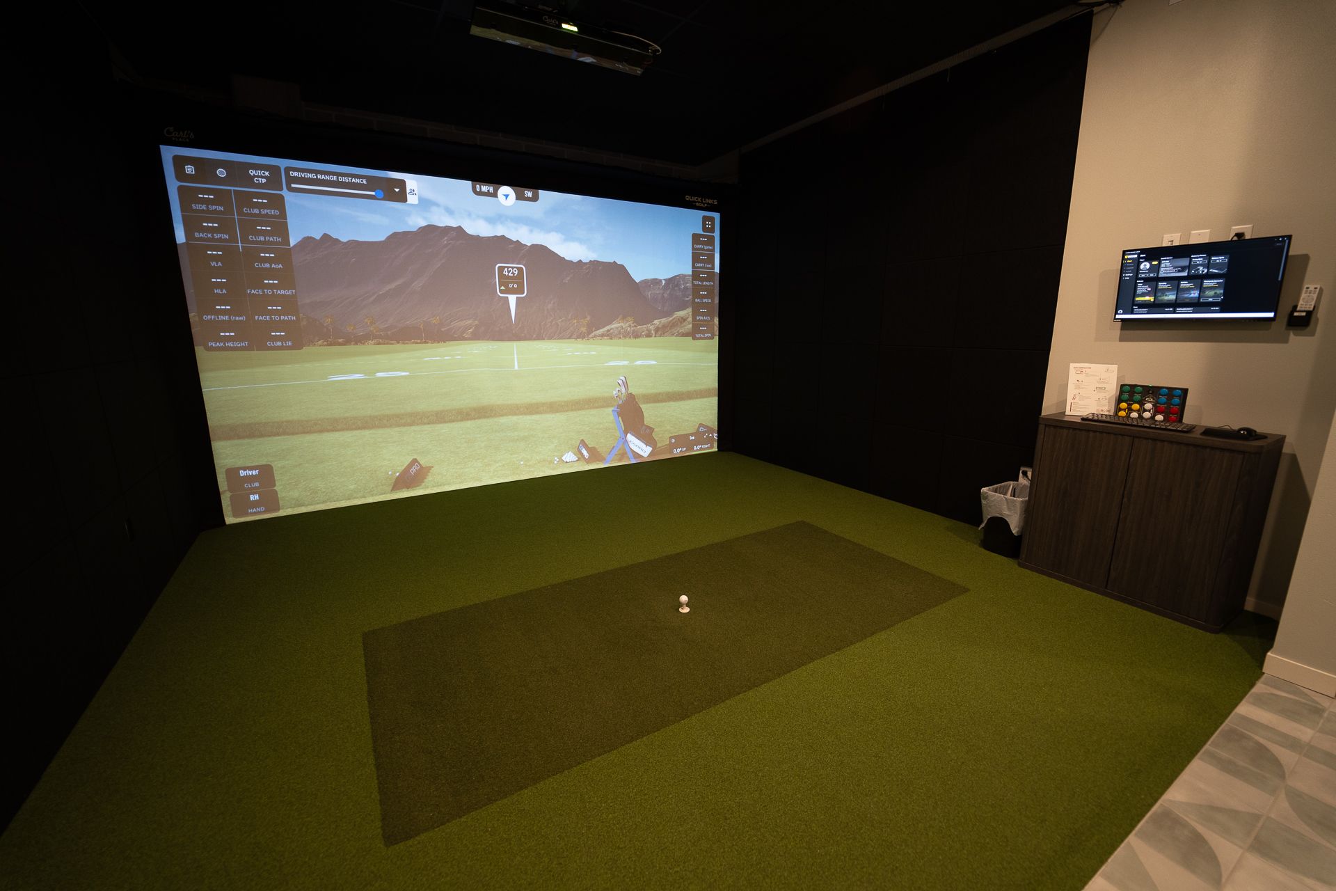 A golf simulator room featuring a large screen displaying a virtual golf course with mountains, a realistic green turf, and a golf ball on a mat. A control panel is on the right. The mood is high-tech and immersive.