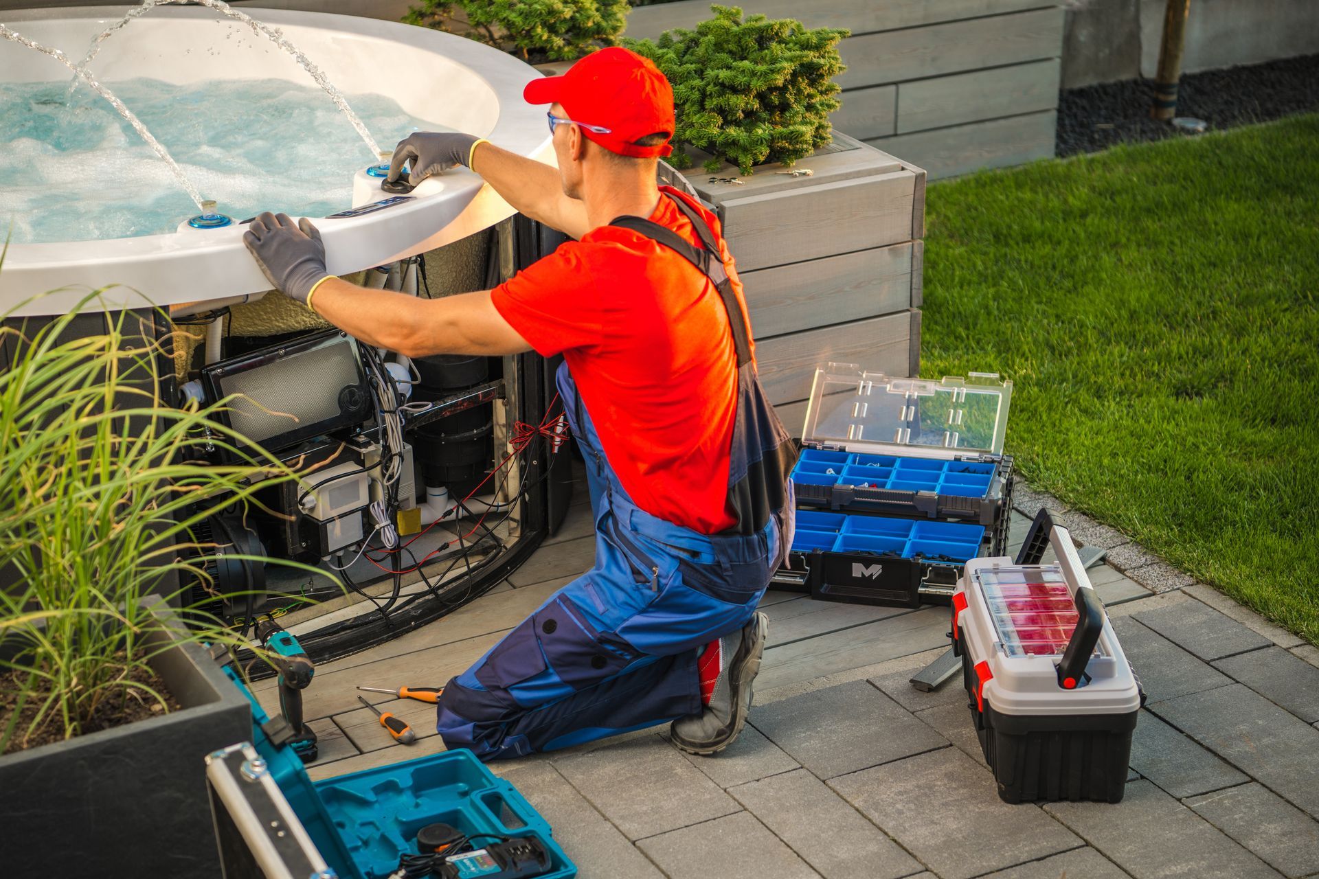 A technician in a red uniform is performing a hot tub repair in a backyard