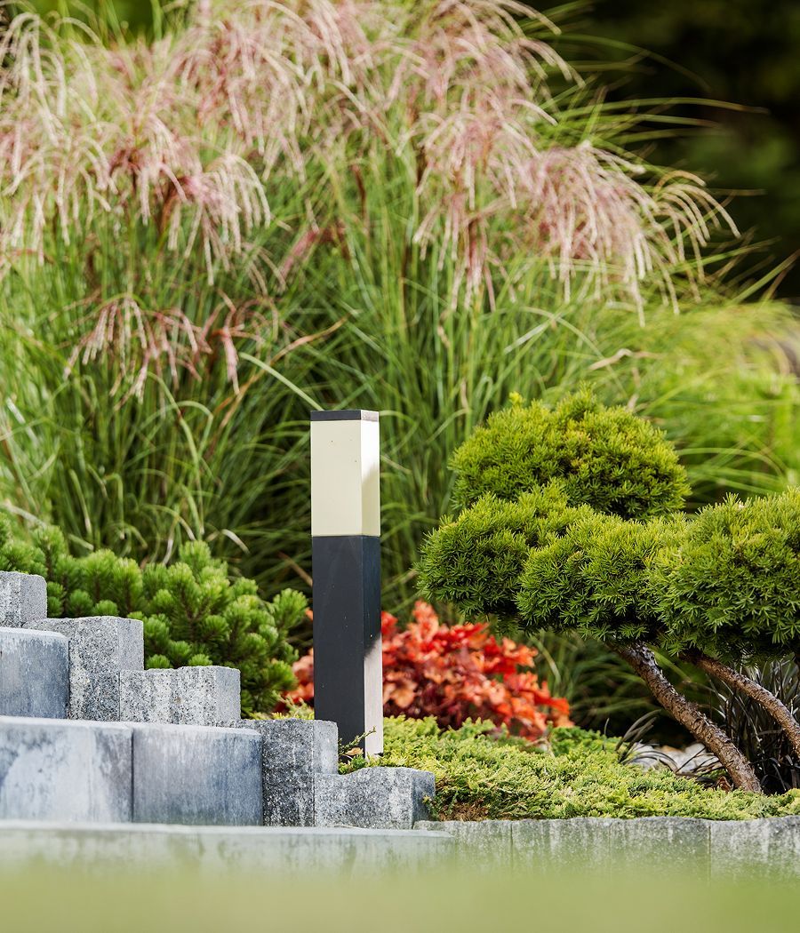 Modern outdoor lamp in a garden setting, surrounded by lush greenery and stone steps.