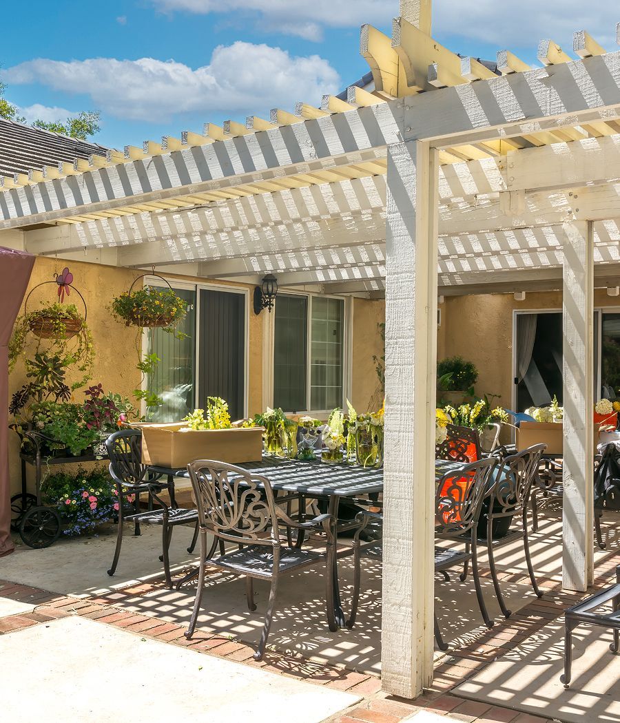 Patio with white pergola, wrought iron furniture, potted plants, and a yellow stucco wall under a blue sky.