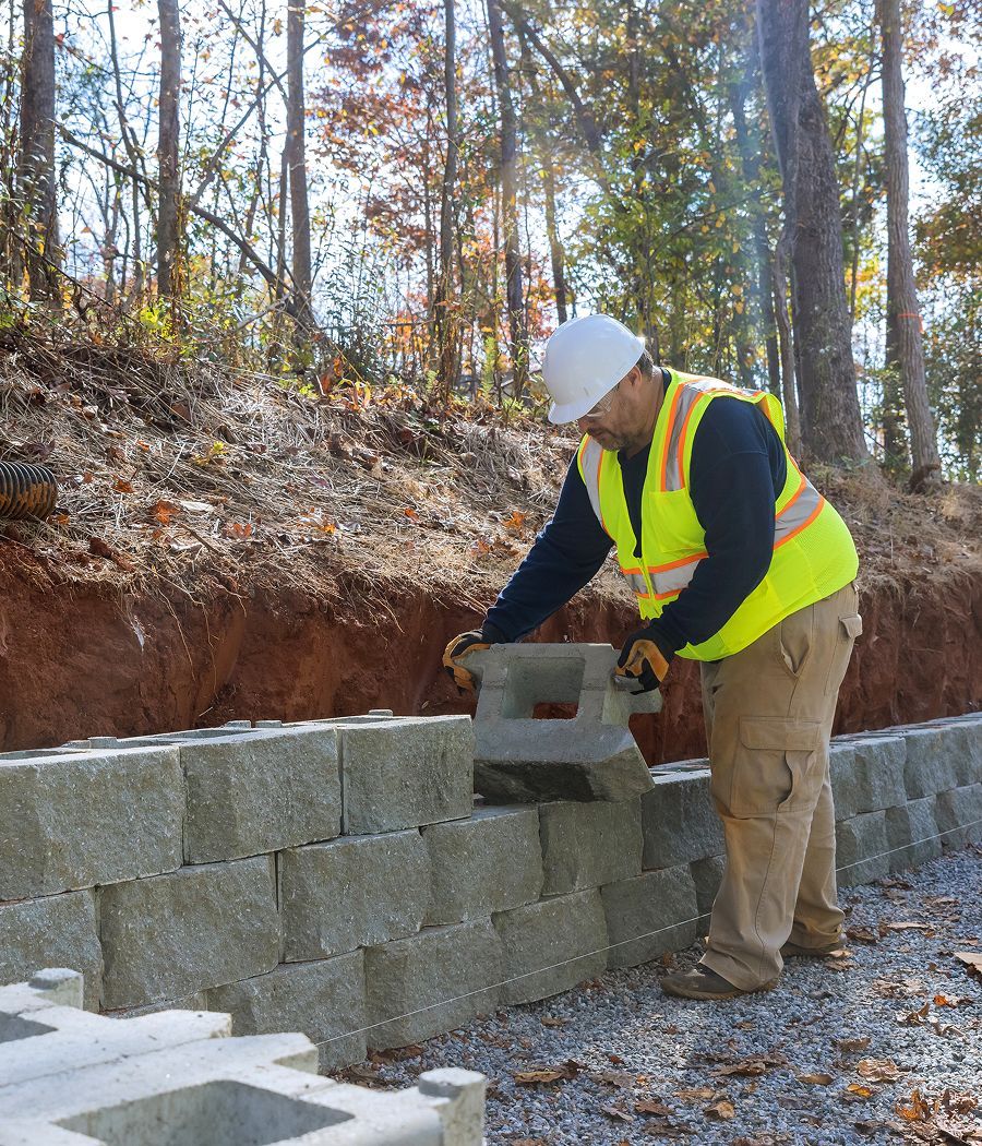 Construction worker in yellow vest and hard hat placing a concrete block on a retaining wall outdoors.