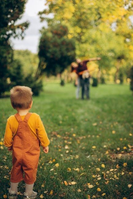 Young child in orange overalls watches blurry figures in a park.