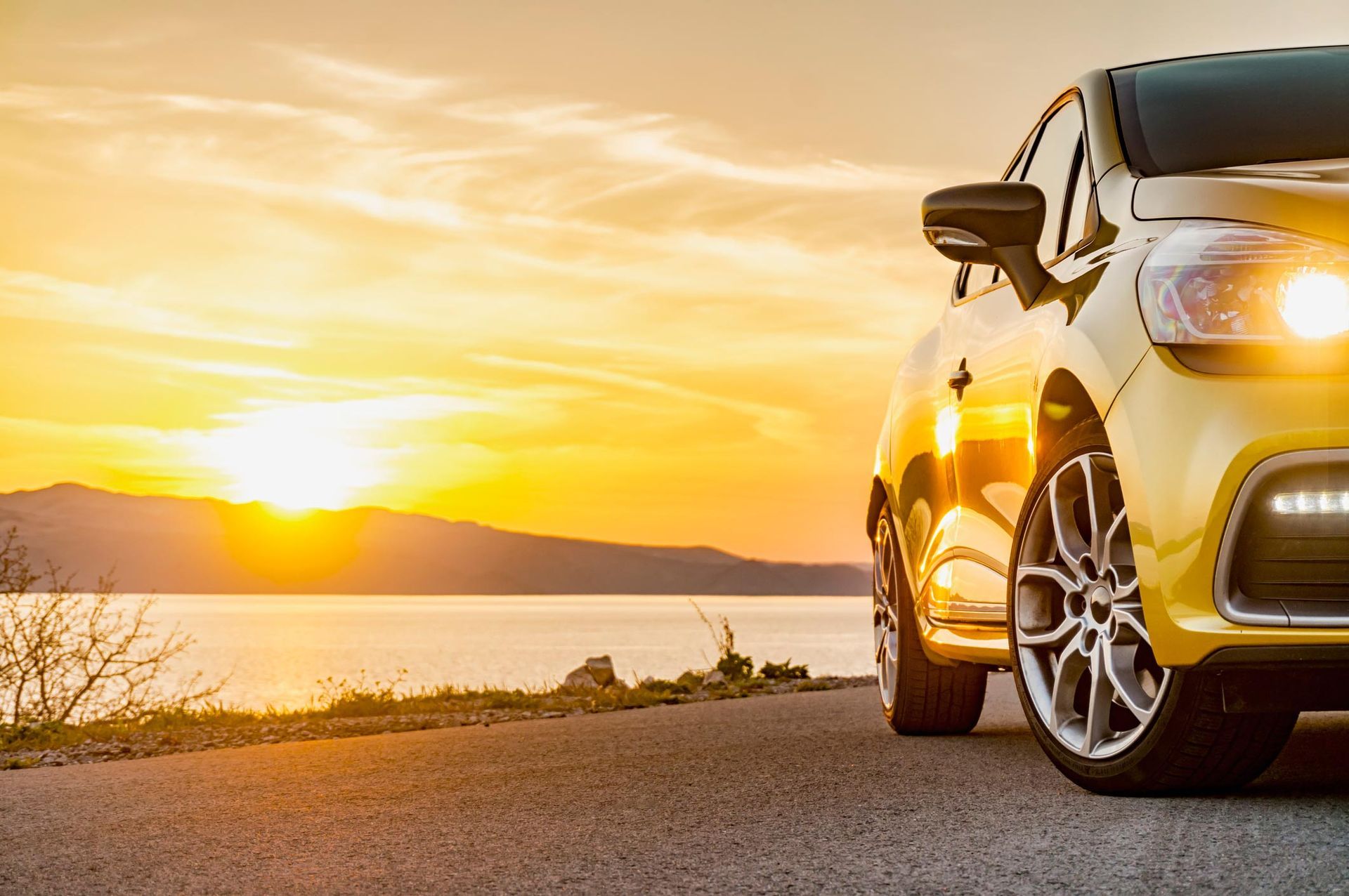 A yellow car is parked on the side of the road next to a body of water at sunset.