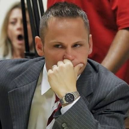 Man in a suit, thoughtful expression, hand near mouth. A woman is in the background with her mouth open.