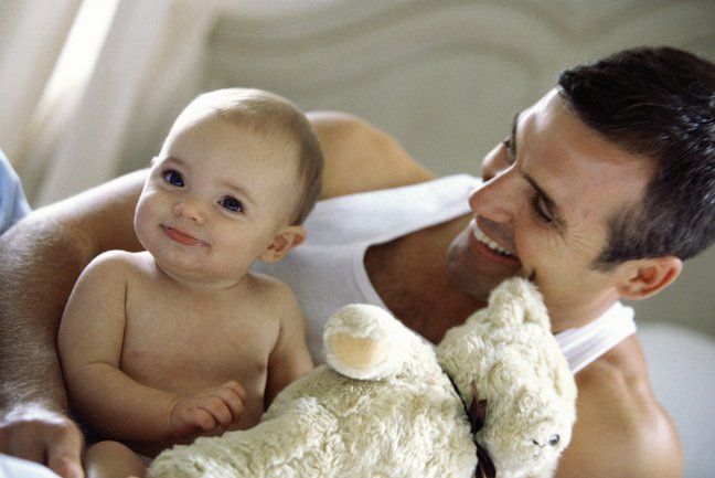 Smiling baby and man holding a teddy bear in a bright setting.