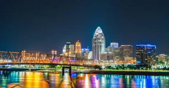 Nighttime view of the Cincinnati skyline across the Ohio River with bridge and city lights reflected in the water.