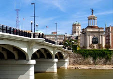 Bridge with arched supports over water, buildings and a clock tower in background, sunny day.
