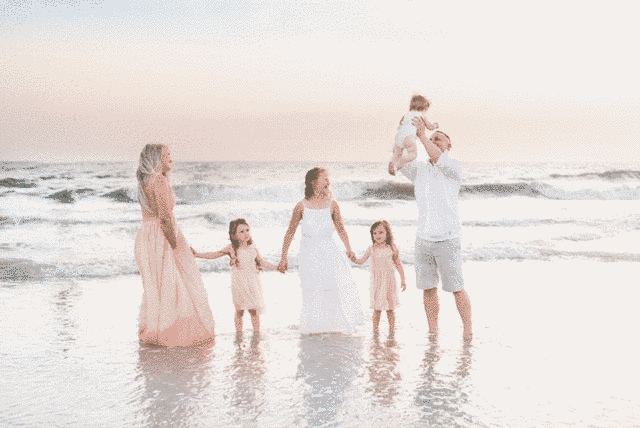 Family of six on beach, two adults holding hands with three children, man holding baby up in air.