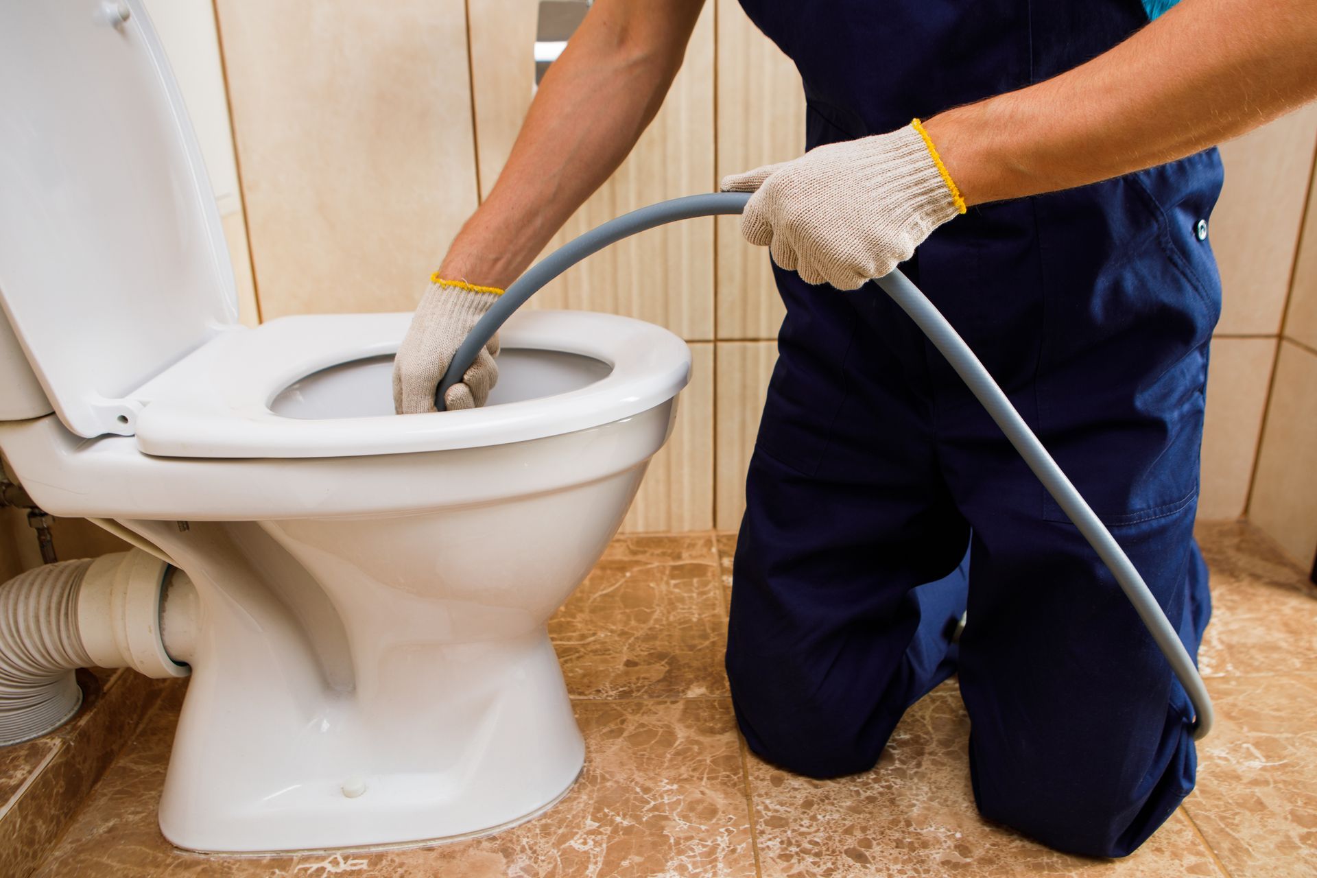 Plumber kneels, inserting a drain snake into a white toilet bowl. He wears gloves and blue overalls.