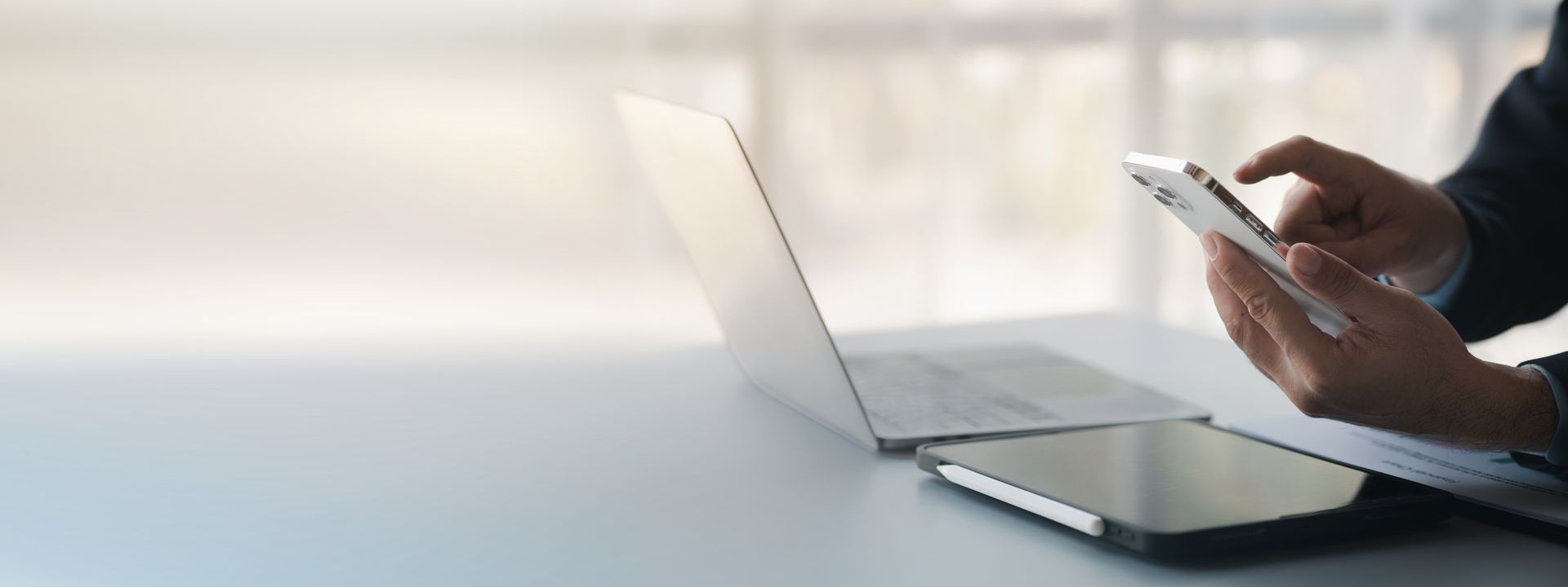 Person using a smartphone at a desk with a laptop and tablet in a bright office.