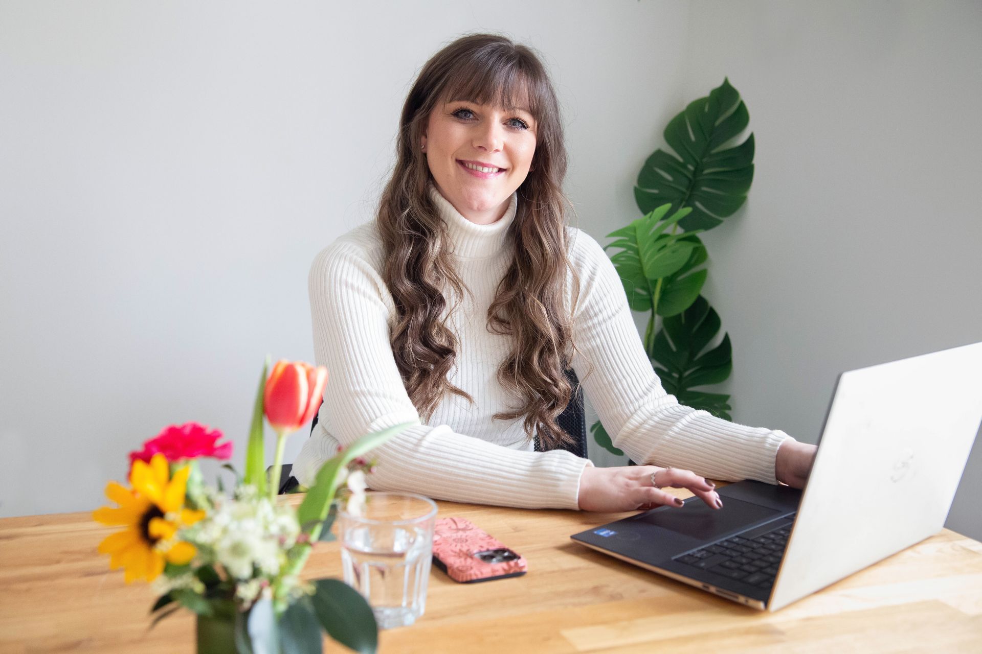 Woman working on a laptop at a wooden desk in an office setting. Papers and plants visible.