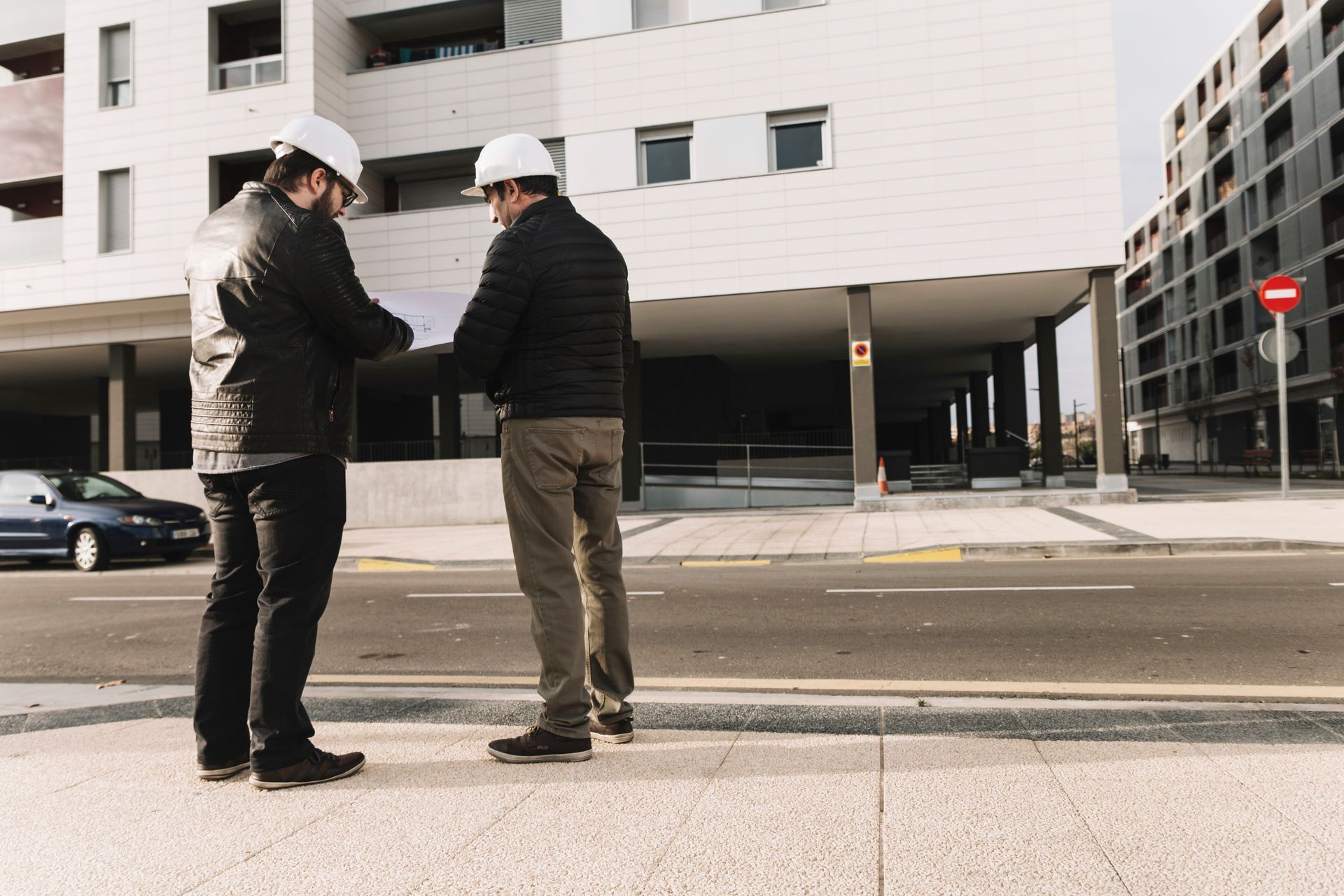 Two men in hard hats reviewing blueprints near a building construction site.
