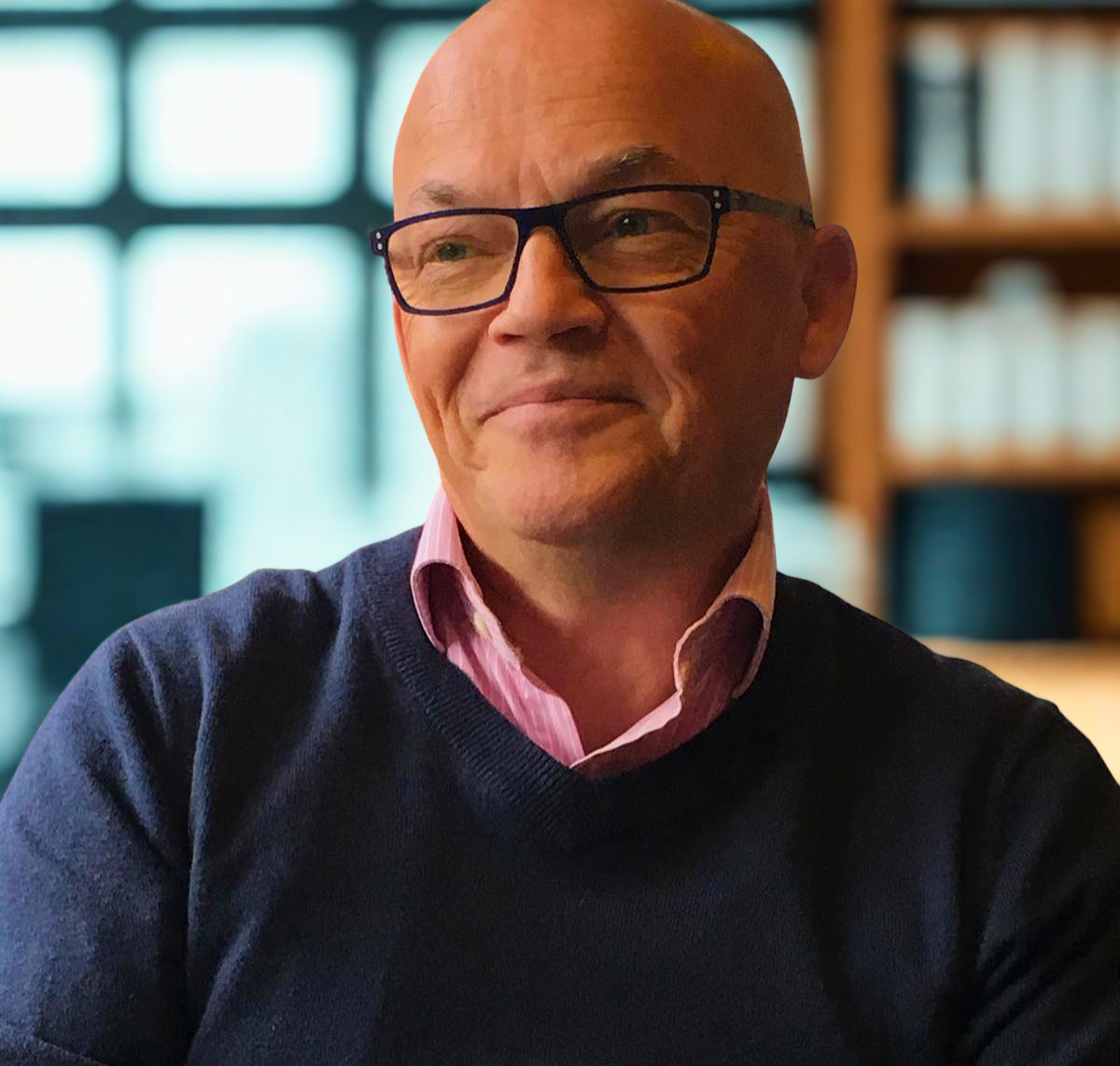 Man with glasses, bald head, wearing a blue sweater and pink collared shirt, smiling, indoors.