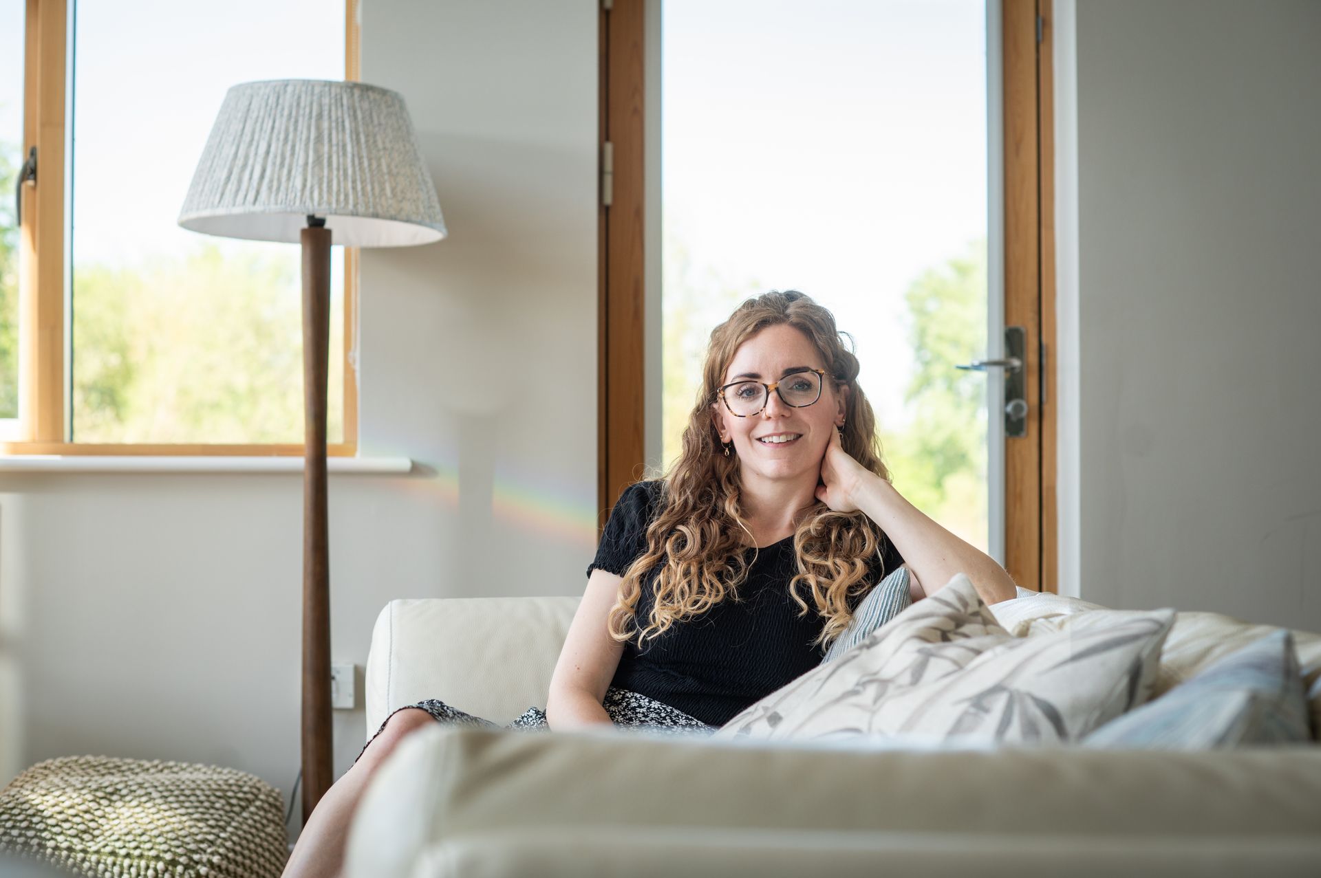 Woman with glasses, sitting on a cream couch, smiles. Beside her is a floor lamp; a window in the background.
