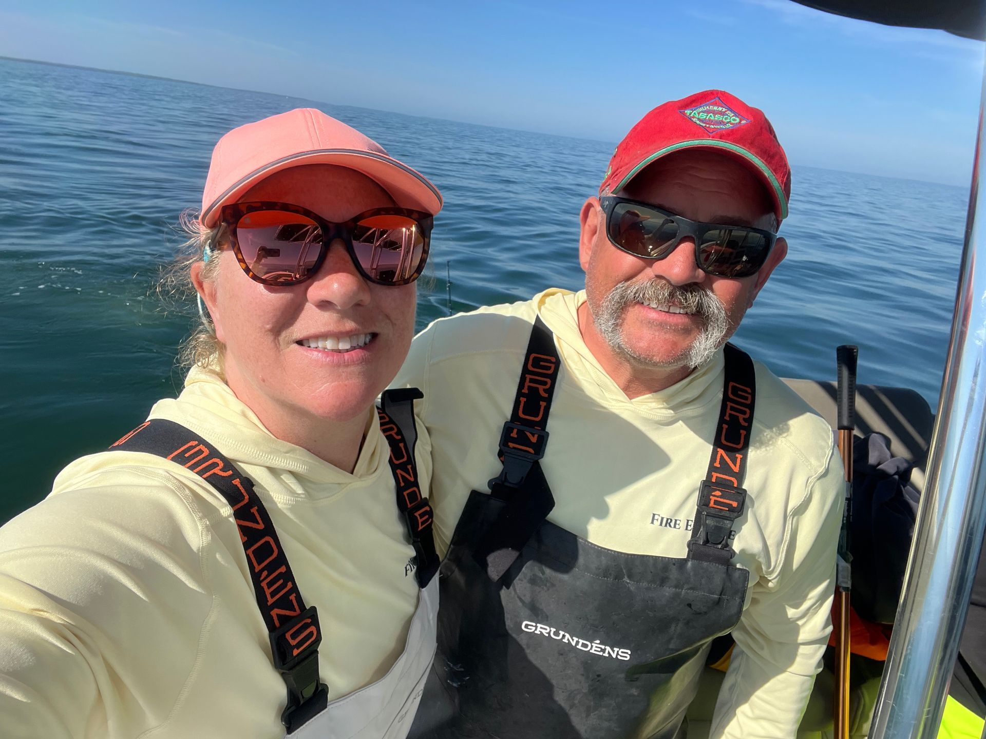 Two people smiling on a boat. Woman in pink cap and sunglasses, man in red cap. Both wearing waders, sea in background.