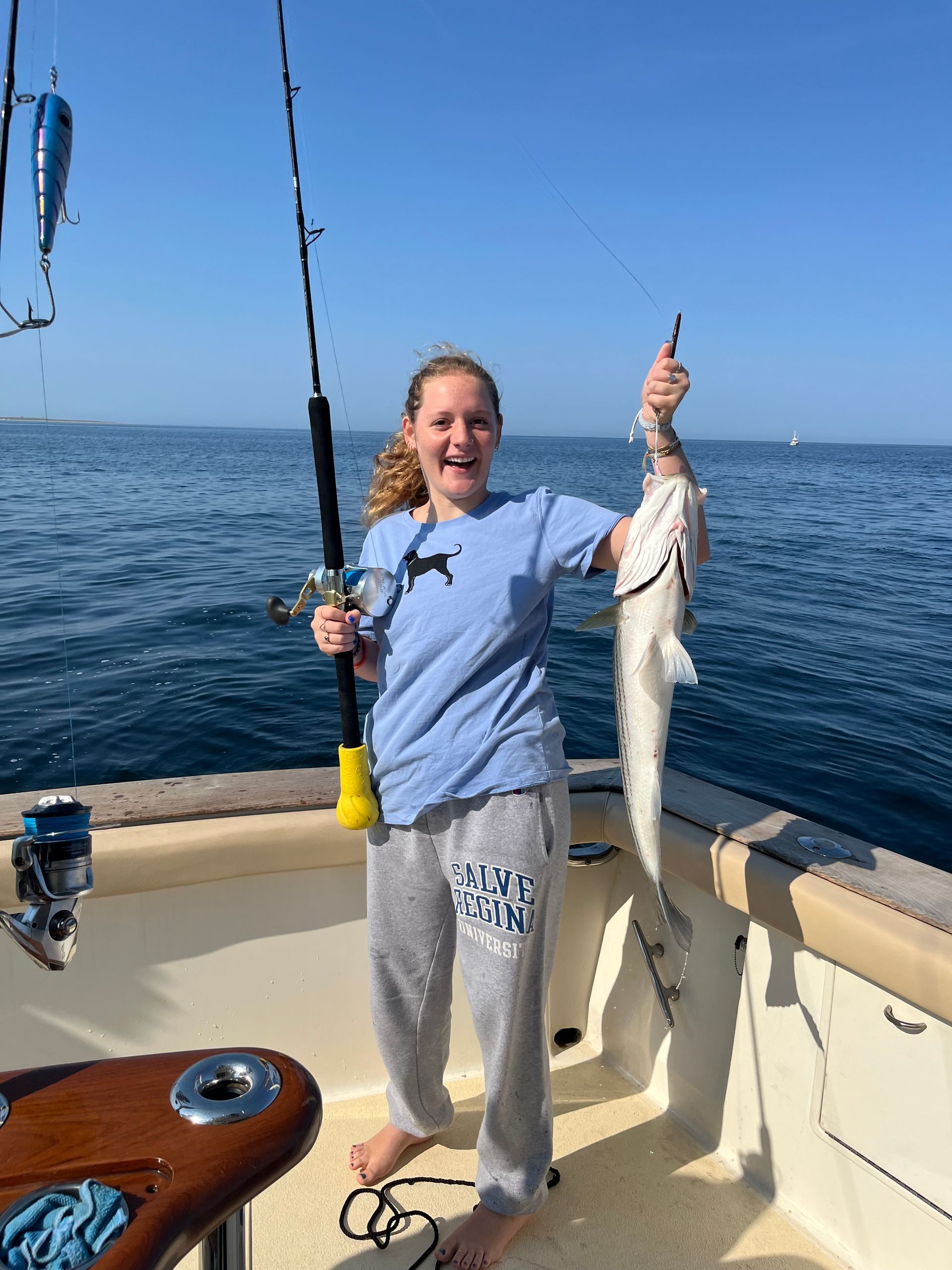 Woman on a boat, holding up a fish she caught, blue sky, water in background.