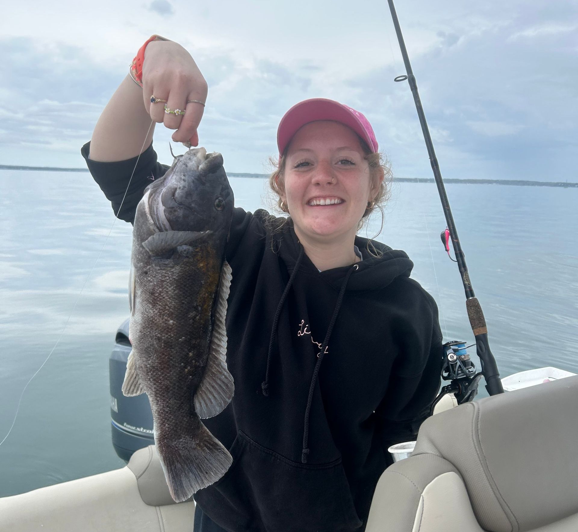 Woman on a boat holding up a black sea bass fish; smiling, ocean in background.